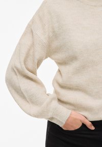 Light beige ribbed knit sweater with long sleeves and a round neckline, featuring a soft texture and relaxed fit. Black pants visible at the bottom.