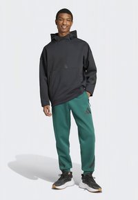 Melnas kapuces džemperis ar priekšējo kabatu, komplektā ar zaļām sporta biksēm ar melnām sānu svītrām. Valkāts ar melniem sporta apaviem.