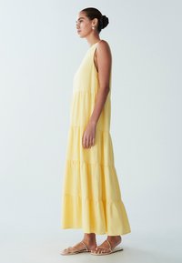 Geltonos daugiapakopės maksi suknelės iš lengvos medžiagos, be rankovių ir su judria siluete. Derinama su minimalistinėmis dirželio formos basutėmis.