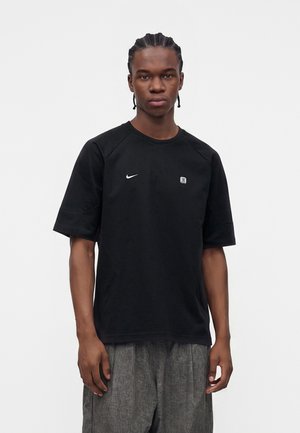 Mladý muž s vrkočmi, ktorý má na sebe čierne tričko Nike a sivé nohavice, stojí pred jednoduchým bielym pozadím.