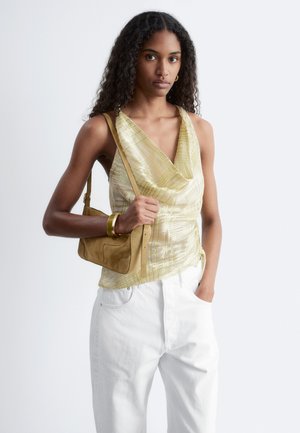 Woman with long curly hair wearing a pale yellow sleeveless top and white pants, holding a tan shoulder bag and wearing a gold bracelet.