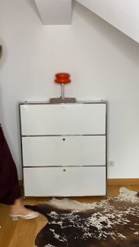 Armário branco de três gavetas com acabamentos em cromo. Uma lâmpada laranja está em cima, ao lado de um tapete com padrão castanho e branco sobre um chão de madeira.