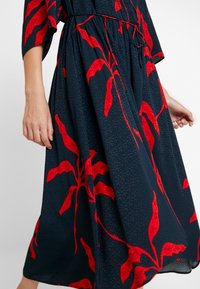 Maxi šaty z tmavě modré látky s texturovaným vzorem, s velkými motivy červených listů; volný střih s nařaseným pasem a širokými rukávy.