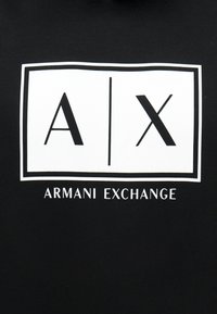 Μαύρο ύφασμα με λευκό ορθογώνιο λογότυπο που περιέχει τα γράμματα "A" και "X" διαχωρισμένα με μια κάθετη γραμμή, και το κείμενο "Armani Exchange" από κάτω.