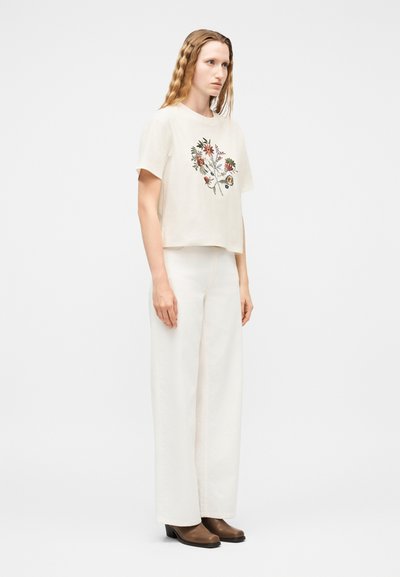 Γυναίκα με μακριά κυματιστά μαλλιά που φοράει κρεμ κοντομάνικη μπλούζα με κεντημένα λουλούδια, λευκό παντελόνι με φαρδιά πόδια και καφέ μπότες, όρθια.