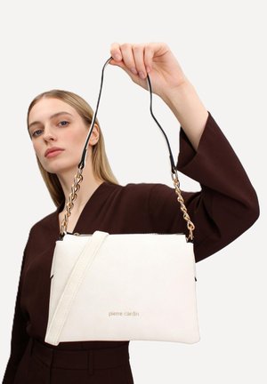 Femme en tenue sombre tenant un sac à main blanc Pierre Cardin avec chaîne dorée et bandoulière en cuir sur fond uni.