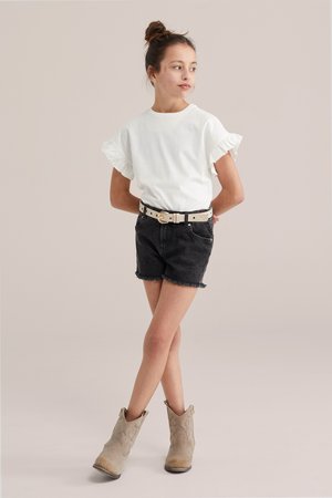 Fată tânără stând cu mâinile la spate, purtând un top alb cu mâneci cu volane, pantaloni scurți din denim negru cu curea și cizme scurte bej.