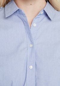 Jasnoniebieska koszula z długim rękawem i wskazującym kołnierzykiem, z białymi guzikami i subtelną teksturą, prezentująca staranne szwy wzdłuż pliski.