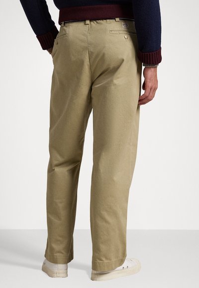 Chinos kaki avec une coupe droite, poches arrière et couture visible. Portés avec des baskets beiges, avec une texture douce semblable au coton.