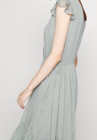 Kvinna i en ljusblå ärmlös klänning med metalliska vertikala ränder och samlad midja, visad från sidan mot en enfärgad bakgrund.