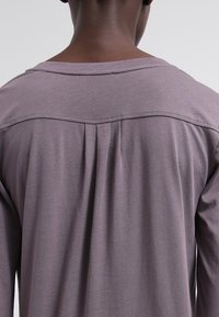 Vista posteriore di una persona che indossa una camicia viola lunga e ampia con maniche lunghe, dotata di un giogo cucito e di un pieghe centrale sulla parte superiore della schiena.