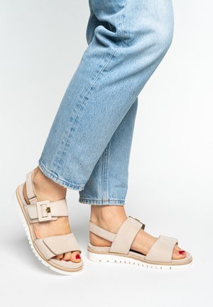 Beige Wildledersandalen mit zwei breiten Riemen, einer Schnallendetail und einer weißen, strukturierten Gummisohle; kombiniert mit hochgekrempelten hellblauen Jeans.
