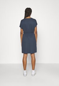 Dunkelblaues Kleid mit kurzen Ärmeln, gerafftem Bund und knielangem Saum. Aus weichem Stoff; kombiniert mit weißen Sneakers.