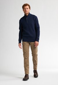 Pull en tricot marine avec un col haut et deux boutons, accompagné d'un pantalon cargo beige et de bottes à lacets marron. L'ensemble a un style décontracté.