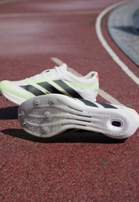 Chaussures de course légères avec une tige blanche et vert néon, des rayures noires, et une semelle blanche texturée avec des pointes argentées pour l'adhérence.