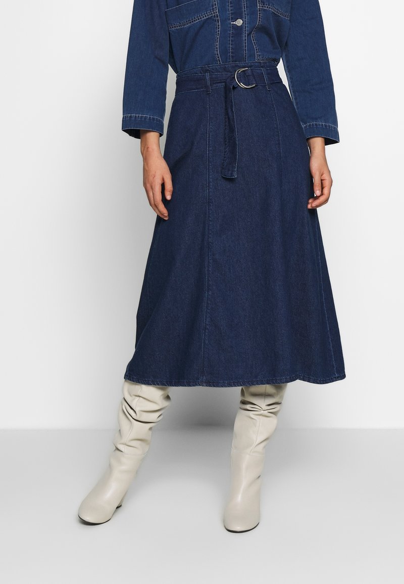 Falda de mezclilla en azul oscuro, diseño en línea A con cintura con cinturón, que presenta una textura suave y detalles de costura visibles. Combinada con botas de rodilla en color crema.