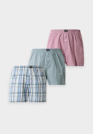 Jeune homme debout pieds nus portant un boxer vert clair sur un fond blanc uni, les mains derrière le dos.