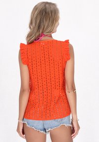 Top arancione con occhielli e maniche a volant, caratterizzato da pattern a intaglio. Abbinato a pantaloni shorts in denim, che mostrano un design casual e traspirante.