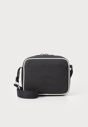 Bolsa crossbody preta texturizada com fecho de zip e acabamento em branco em contraste. Apresenta o logotipo em relevo da Lacoste na frente e uma alça ajustável.