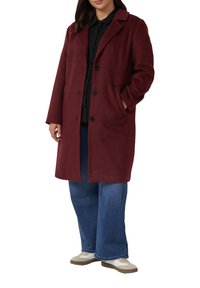 Bordeaux wollen blend jas met een geknipte kraag, vier knopen aan de voorkant en zijzakken. Gedragen over een zwarte blouse en blauwe wijde jeans.