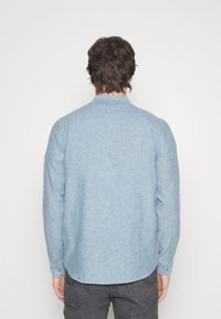 Chemise à manches longues bleu clair avec un col mandarin, en tissu texturé, dotée de poignets boutonnés et d'un ourlet incurvé à l'arrière.