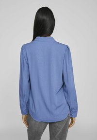 Lange mouwen shirt van lichtblauwe stof. Heeft een subtiel stippenpatroon, een klassieke kraag en knopenmanchetten. De achterkant heeft een afgeronde onderkant.