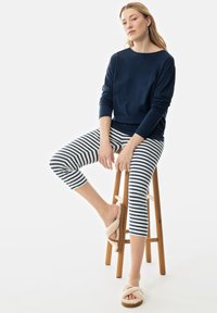 Marineblauw langemouwtop, gestreepte leggings in het wit en marineblauw, en luchtige beige slippers. Model zit op een houten kruk. Eenvoudig, casual ontwerp.