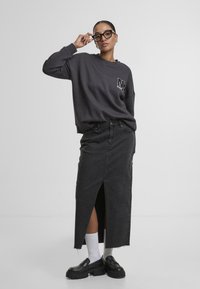 Sweatshirt ample gris foncé avec un logo sur la poitrine, associé à une jupe en denim noire à fente haute et des mocassins noirs épais. Chaussettes blanches visibles.
