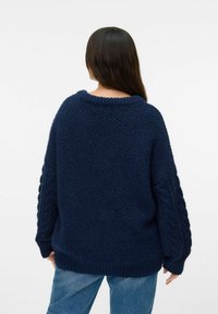 Pull en tricot bleu marine avec une texture épaisse, présentant des manches larges en tricot torsadé et un col et un ourlet côtelés. Vue arrière présentée.