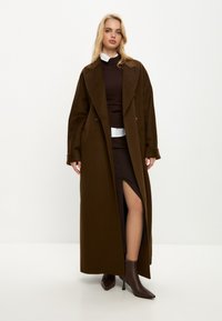 Brauner Wollmischungs-Mantel mit breitem Kragen, doppelter Knopfleiste und langen Ärmeln; kombiniert mit einem dunklen Kleid und spitzen Absatzstiefeln.