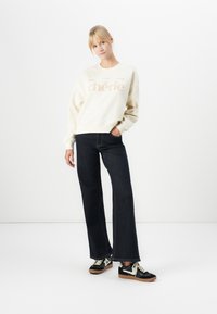 Mujer de pie con la mano en el bolsillo, vistiendo un sudadera color crema con el texto "chérie", jeans oscuros de pierna ancha y zapatillas negras y blancas.