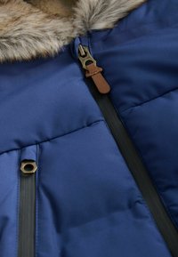 Blaue gepolsterte Jacke mit Kunstfellkragen, sichtbaren schwarzen wasserdichten Reißverschlüssen und einem Reißverschlussanhänger aus braunem Leder.