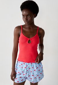 Top rosso senza maniche con dettaglio a chiave, abbinato a pantaloni corti azzurri chiari con un motivo a ciliegie e una stampa a quadri.