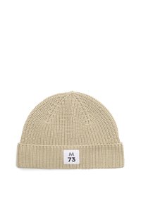 Beige kötött sapka hajtott széllel és fehér címkével, amelyen fekete "M 73" felirat található az elülső középen.