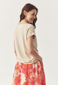 Beige top s krátkým rukávem a ohrnutými manžetami, spárovaný s květinovou sukní v živých červených a oranžových barvách. Měkká textura, volný střih.