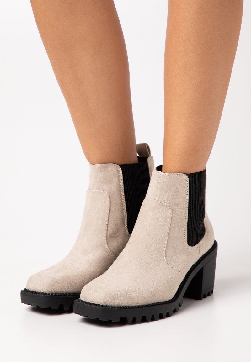 Bottines beige en daim avec panneaux latéraux élastiques noirs et talons noirs épais portées sur jambes nues.