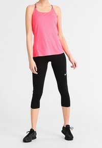 Una donna indossa una canotta sportiva rosa senza maniche, leggings neri a tre quarti e scarpe da corsa nere, mentre si trova in una posa rilassata su uno sfondo bianco.