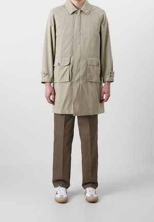 Personne portant un manteau beige mi-long à boutons sur les poches, un pantalon marron et des baskets blanches avec des accents rouges, debout devant un fond uni.