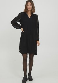Schwarzes, plissiertes Kleid mit durchsichtigen langen Ärmeln, V-Ausschnitt und ausgestelltem Schnitt, kombiniert mit schwarzen Leder-Loafern und blickdichten Strumpfhosen.