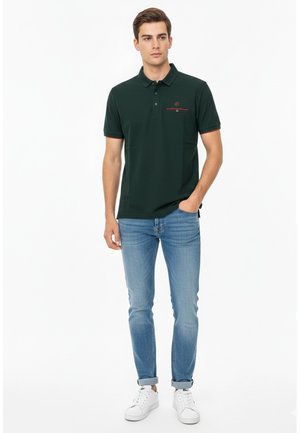 Joven de pie con una mano en el bolsillo, vistiendo una camisa tipo polo verde oscuro, jeans azul claro y zapatillas blancas, sobre un fondo blanco.