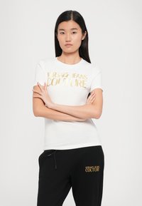 Camiseta blanca con letras doradas "VERSACE JEANS COUTURE", combinada con pantalones de chándal negros que presentan bordados dorados similares. Acabado de tejido suave.