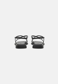 Zwarte platte sandalen hebben kruisende banden, een gladde matte textuur, een rechthoekige zool en een minimalistisch ontwerp zonder versieringen.