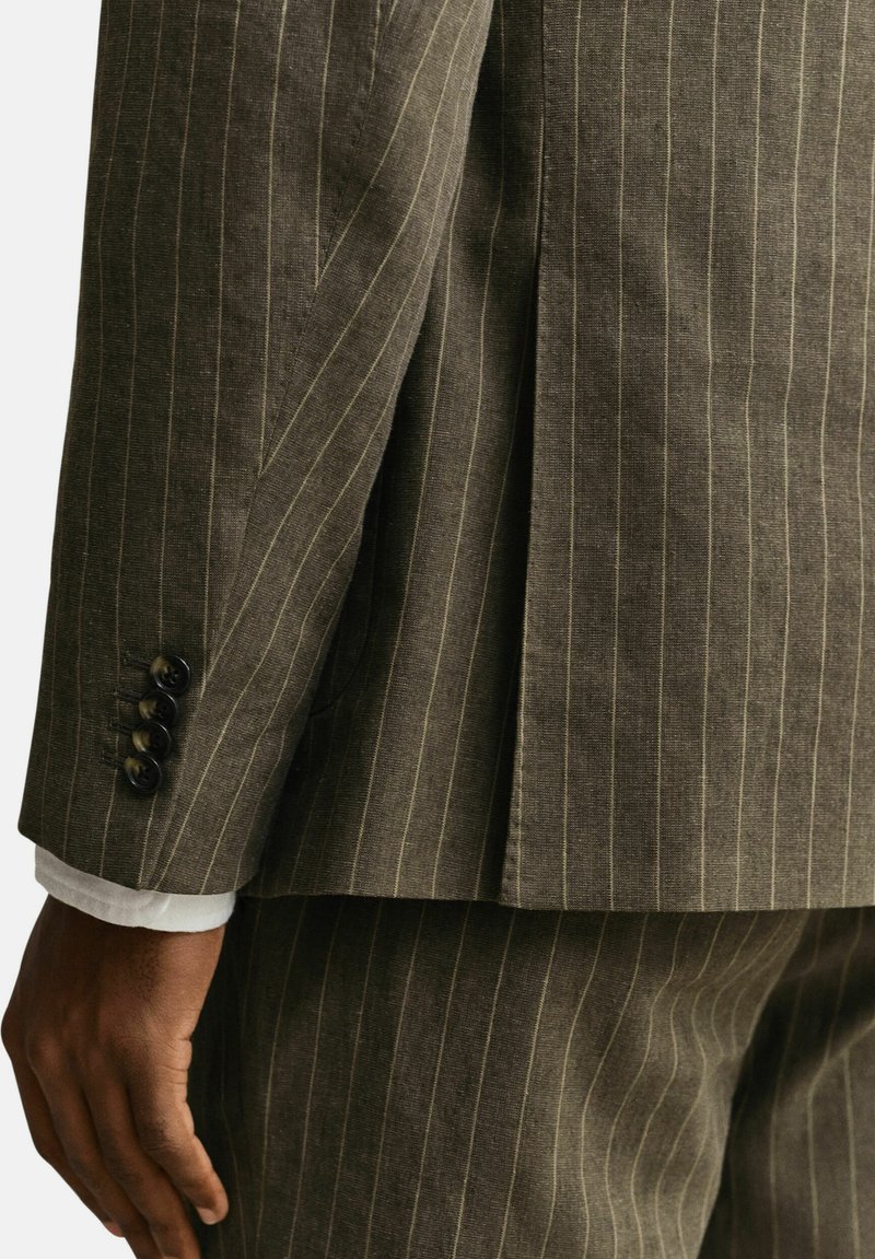 Bruine krijtstreep pakjas en broek gedragen door een persoon met een donkere huid, met zicht op de knopen van de jas en de witte manchetten van het overhemd.