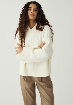 Femme aux longs cheveux bouclés portant un pull blanc oversize et un pantalon marron, debout, les bras croisés, sur un fond uni.