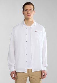 Camicia bianca in lino con taschino e un piccolo dettaglio di bandiera ricamato. Si adatta in modo morbido, con maniche lunghe e chiusura a bottoni.