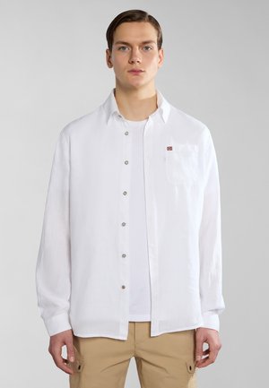 Camicia bianca in lino con taschino e un piccolo dettaglio di bandiera ricamato. Si adatta in modo morbido, con maniche lunghe e chiusura a bottoni.