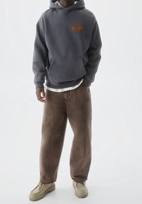 Grijze capuchontrui met voorzak en oranje logo, gecombineerd met losse bruine denim broek en beige schoenen. Simpele, casual stijl.