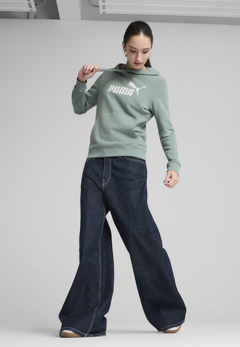 Felpa verde chiaro con logo Puma bianco e tasca frontale, abbinata a jeans in denim scuro a gamba larga con cuciture in vista e vestibilità comoda.