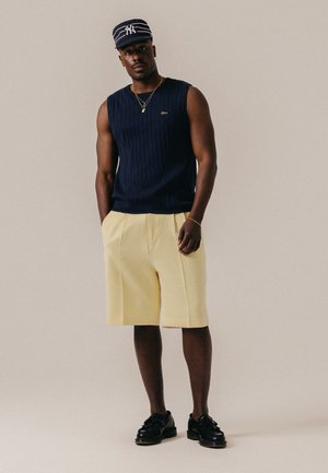 Mand iført navy ærmeløs sweater, creme knælange shorts, sorte sko og navy stribet kasket stående mod ensfarvet baggrund.