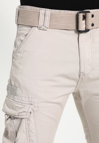 Béžové cargo kalhoty s texturovanou tkaninou, vybavené širokým tkaným páskem, bočními kapsami a nášivkou loga na zadní kapse.